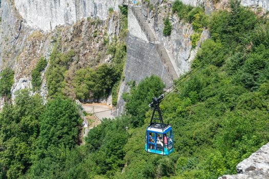 Dinant, Belgium - June 26, 2019: Blue cable car against green foliage bringing visitors up along gray stone cliffs and ramparts of Citadelle.