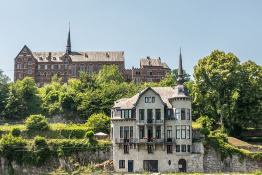 Dinant, Belgium - June 26, 2019: Red stone ancient convent of the Capuchins, now hotel, museum and administratif building and Villa Mouchenne, top restaurant, in front. Blue sky, green foliage.