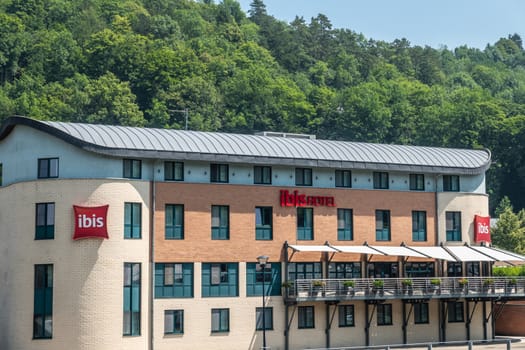 Dinant, Belgium - June 26, 2019: Beige and red stone building of Ibis Hotel along Meuse River backed by green foliage.