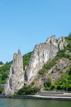 Dinant, Belgium - June 26, 2019: South side of Le rocher Bayard along La Meuse River surrounded by green foliage under blue sky.