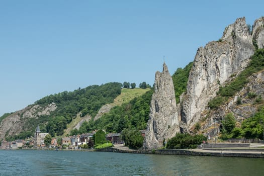 Dinant, Belgium - June 26, 2019: Wider view on South side of Le rocher Bayard along La Meuse River surrounded by green foliage under blue sky. Saint Paul church in distance.