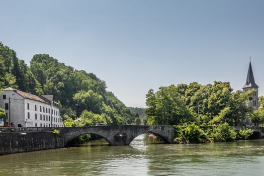 Dinant, Belgium - June 26, 2019: Old roman bridge over greenish Lesse River. Saint Anne Church tower captured in green foliage on the right. White building on the left under light blue sky.