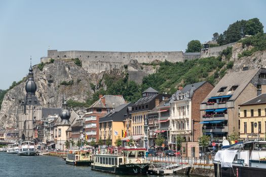 Dinant, Belgium - June 26, 2019: Citadelle fortification seen from right south bank of Meuse River with church and buildings, cars, boats under blue sky.