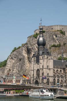 Dinant, Belgium - June 26, 2019: Notre Dame church on River Meuse bank at Charles De Gaule bridge at bottom of Citadelle. Boat on river.