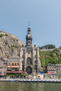 Dinant, Belgium - June 26, 2019: Frontal view on gray stone Notre Dame church from Meuse River with Citadelle fort ramparts in back under blue sky, some green foliage.