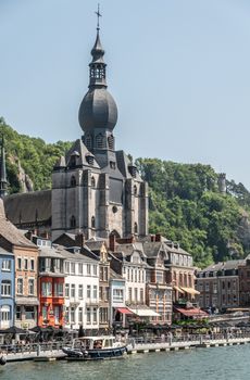 Dinant, Belgium - June 26, 2019: Notre Dame church on River Meuse north right bank at Charles De Gaule bridge behind businesses and restaurants under blue sky. Green foliage.