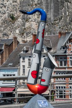 Dinant, Belgium - June 26, 2019: Red, gray and blue saxophone statue to honor the European Union on Charles de Gaulle bridge. Business and houses in back against gray rock cliff.