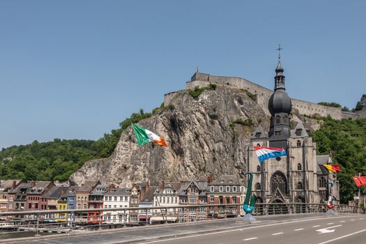 Dinant, Belgium - June 26, 2019: Notre Dame church and Citadelle from Charles De Gaule bridge with businesses on right north bank of Meuse River, under blue sky. Green foliage.