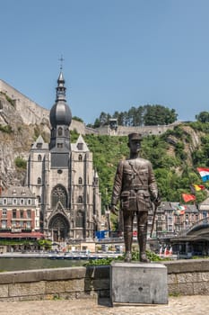 Dinant, Belgium - June 26, 2019: Notre Dame church and Charles De Gaule statue on opposite banks of Meuse River, under blue sky. Green foliage. Multiple flags.