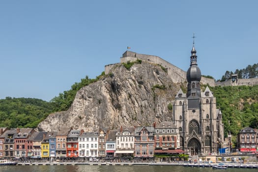 Dinant, Belgium - June 26, 2019: Notre Dame church and Citadelle from other river Meuse bank with businesses on right north bank, under blue sky. Green foliage.