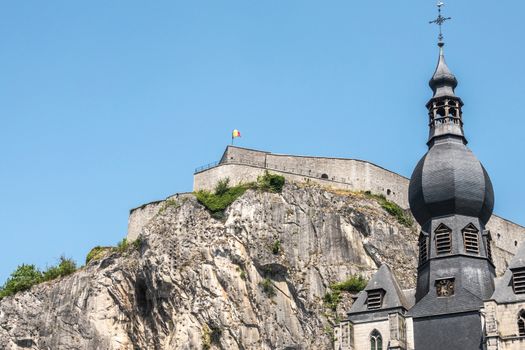 Dinant, Belgium - June 26, 2019: Closeup of Citadelle front top with Belgian flag and spire of Notre Dame church against blue sky.