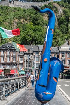 Dinant, Belgium - June 26, 2019: blue saxophone statue on Charles de Gaulle bridge. Business and houses in back. Country flags and people.