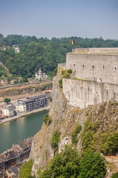 Dinant, Belgium - June 26, 2019: tip of Gray stone south ramparts of Citadelle fortification with Belgian Flag at top under blue sky. Horizon is forest and down the Meuse River and cityscape.