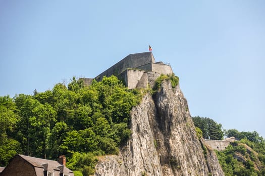 Dinant, Belgium - June 26, 2019: Closeup of fish eye view of Citadelle front top with Belgian flag under blue sky and green foliage.