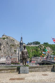 Dinant, Belgium - June 26, 2019: Notre Dame church, Citadelle and Charles De Gaule statue on opposite banks of Meuse River, under blue sky. Green foliage. Multiple flags.