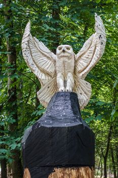 Han-sur-Lesse, Belgium - June 25, 2019: Animal park with Snowy owl statue in closeup, on blackened tree stump with green foliage in back.