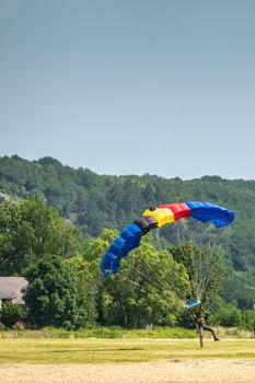 Han-sur-Lesse, Belgium - June 25, 2019: Belgian soldier on parachute with Belgian flag colors lands on field with forested hills and blue sky in back.