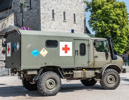 Han-sur-Lesse, Belgium - June 25, 2019: Belgian army green ambulance truck with red cross symbol downtown and green foliage. Grau stone building in back.