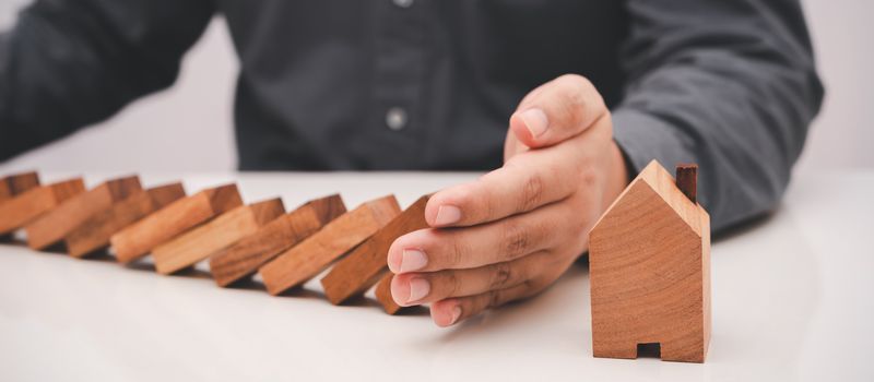 business man hand prevent falling wooden stick (dominoes) to the house model . risk management in business concept.