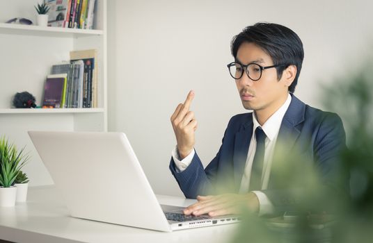 Angry or Irritable Asian Businessman Wear Eyeglasses in Formal Navy Blue Suit Show Middle Finger in front of Laptop Screen. Angry Asian businessman serious working in office