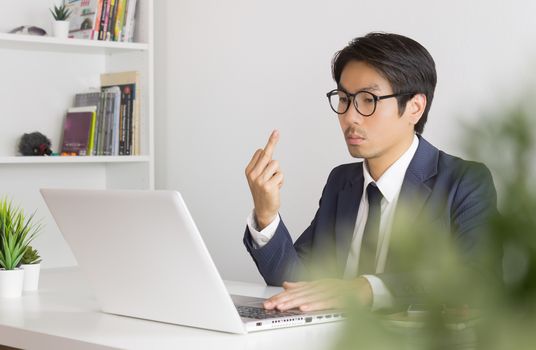 Angry or Irritable Asian Businessman Wear Eyeglasses in Formal Navy Blue Suit Show Middle Finger in front of Laptop Screen. Angry Asian businessman serious working in office