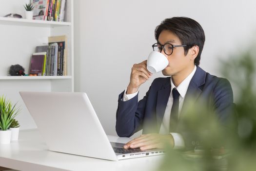 Asian Businessman Wear Eyeglasses and Drink Coffee in front of Laptop. Relax between working time of Asian businessman