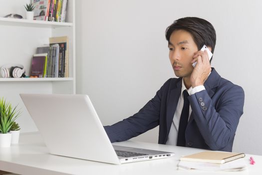 Asian Financial Advisor or Asian Consulting Businessman in Suit Talking with Customer by use Smartphone and Laptop. Asian Financial Advisor or Asian Consulting Businessman working in office