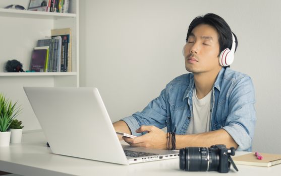 Portrait Asian Photographer with Laptop and Smartphone and Headphone in Home Office. Photographer listening to music by use bluetooth technology in smartphone