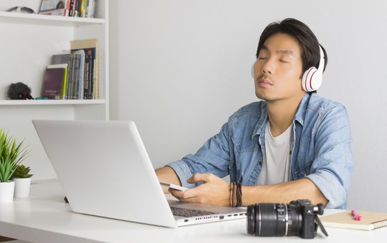 Portrait Asian Photographer with Laptop and Smartphone and Headphone in Home Office. Photographer listening to music by use bluetooth technology in smartphone