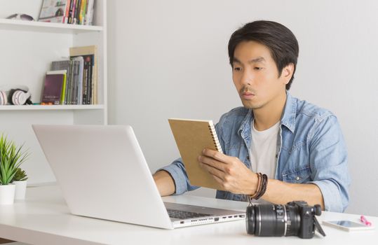 Asian Photographer or Freelancer in Denim or Jeans Shirt Checking Work Schedule in Notebook in front of laptop in Home Office. Photographer or freelancer working with technology