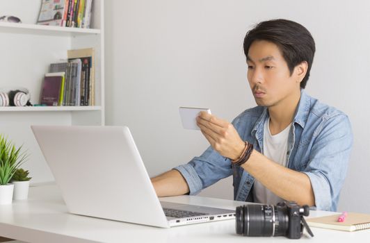 Asian Photographer or Freelancer in Denim or Jeans Shirt Checking Photo File by Smartphone in front of Laptop in Home Office. Photographer or freelancer working with technology
