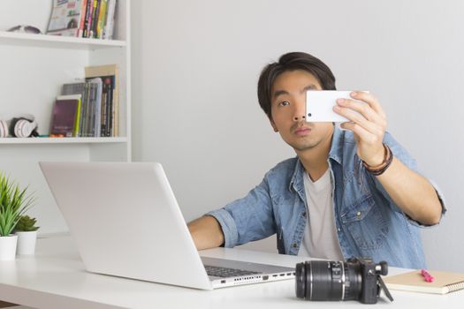 Asian Photographer or Freelancer in Denim or Jeans Shirt Selfie or Take Photo by Smartphone in Front of Laptop in Home Office. Photographer or freelancer working with technology