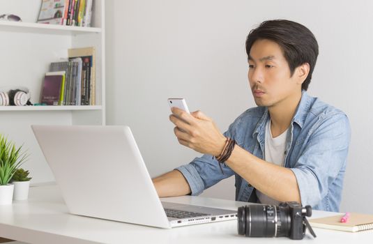 Asian Photographer or Freelancer in Denim or Jeans Shirt Send Message or Chat with Colleague by Smartphone in front of Laptop in Home Office. Photographer or freelancer working with technology