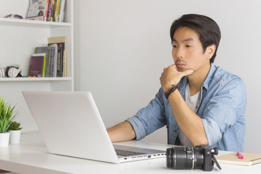 Asian Photographer or Freelancer in Denim or Jeans Shirt Serious Checking Photo File in Laptop in Home Office. Photographer or freelancer working with technology