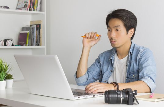 Asian Photographer or Freelancer in Denim or Jeans Shirt Hold Pen and Thinking in Front of Laptop in Home Office. Serious photographer or Freelancer working with technology