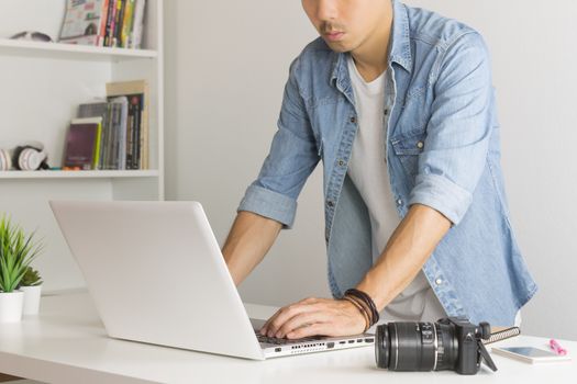 Asian Photographer or Freelancer in Denim or Jeans Shirt Working with Laptop in Standing Posture in Home Office. Photographer or freelancer working with technology