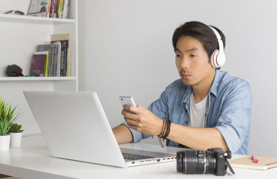 Asian Freelance Videographer in Denim or Jeans Shirt Checking Multimedia File by Smartphone with Laptop in Home Office. Freelance Videographer working with technology