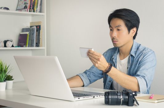 Asian Photographer or Freelancer in Denim or Jeans Shirt Checking Photo File by Smartphone in front of Laptop in Home Office. Photographer or freelancer working with technology