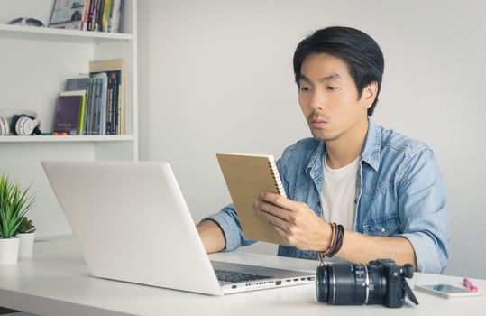 Asian Photographer or Freelancer in Denim or Jeans Shirt Checking Work Schedule in Notebook in front of laptop in Home Office. Photographer or freelancer working with technology