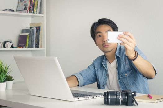 Asian Photographer or Freelancer in Denim or Jeans Shirt Selfie or Take Photo by Smartphone in Front of Laptop in Home Office. Photographer or freelancer working with technology