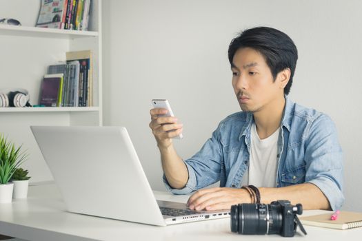 Asian Photographer or Freelancer in Denim or Jeans Shirt Send Message to Customer by Smartphone in front of Laptop in Home Office. Photographer or freelancer working with technology