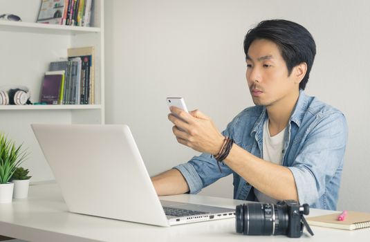 Asian Photographer or Freelancer in Denim or Jeans Shirt Send Message or Chat with Colleague by Smartphone in front of Laptop in Home Office. Photographer or freelancer working with technology