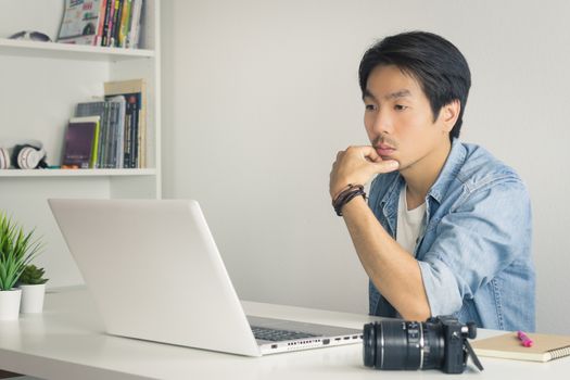 Asian Photographer or Freelancer in Denim or Jeans Shirt Serious Thinking in front of Laptop in Home Office. Photographer or freelancer working with technology