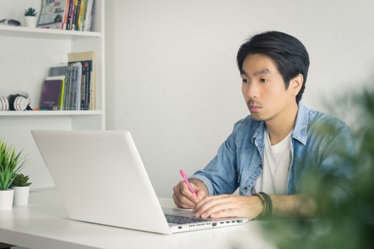 Asian Casual Businessman or Freelancer in Denim or Jeans Shirt Working with Laptop and Note Detail in Home Office. Serious casual businessman or Freelancer working with technology