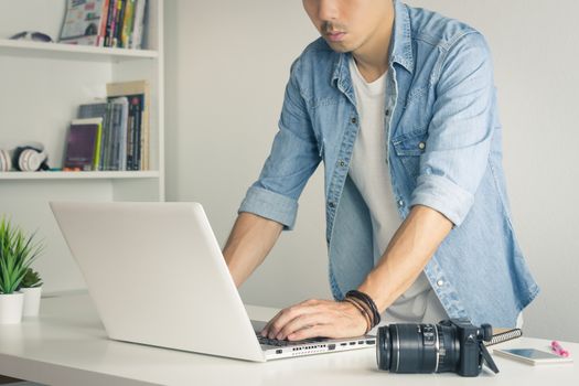 Asian Photographer or Freelancer in Denim or Jeans Shirt Working with Laptop in Standing Posture in Home Office. Photographer or freelancer working with technology