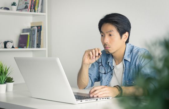 Asian Casual Businessman or Freelancer in Denim or Jeans Shirt Hold Eyeglasses and Working with Laptop in Home Office. Serious casual businessman or Freelancer working with technology