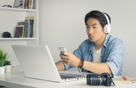 Asian Freelance Videographer in Denim or Jeans Shirt Checking Multimedia File by Smartphone with Laptop in Home Office. Freelance Videographer working with technology