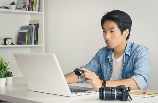 Asian Photographer or Freelancer in Denim or Jeans Shirt Checking Media File in Action Camera with Laptop in Home Office. Photographer or Freelancer working with technology