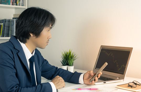 Young Asian Forex Trader or Investor or Businessman in Suit Hold Smartphone and Trading Forex or Stock Chart by Laptop in Trader Room  in Vintage Tone