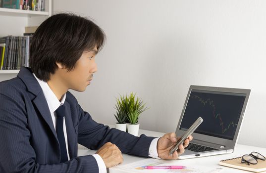 Young Asian Forex Trader or Investor or Businessman in Suit Hold Smartphone and Trading Forex or Stock Chart by Laptop in Trader Room